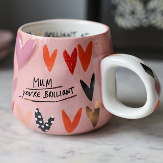 Lifestyle image of the Mum, You're Brilliant Mug displayed on a white marble counter with kitchen accessories in the background. 