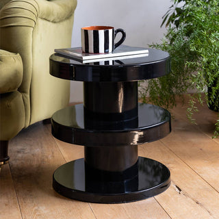 The Glossy Black Side Table styled with a mug and magazine on the top, displayed next to a sofa and a plant on a wooden floor. 