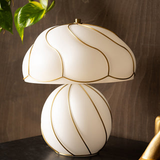 The Beautiful Mushroom Hayworth Table Lamp displayed on a black table in front of a dark wood wall, with a plant and candle holder also in the shot.