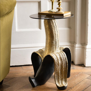 The Charcoal & Gold Banana Side Table displayed next to a sofa in front of a white panelled wall, with a book and wax-covered candlesticks on the top.