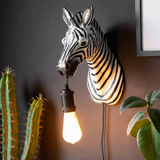 The Zebra Wall Light switched on and styled on a black wall, next to a framed print and some plants.