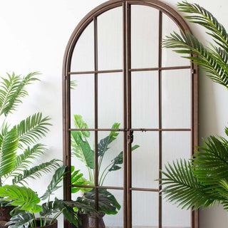 Image of Rockett St George Black Metal Arch Window Pane Indoor/Outdoor Mirror With Opening Doors displayed on a wall surrounded by home accessories and foliage. 