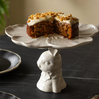 The Decorative Rabbit Cake Plate displayed with a carrot cake on top, surrounded by kitchen accessories on a black table.