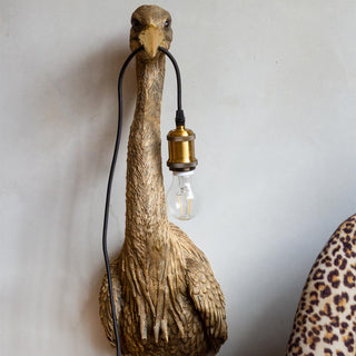 Lifestyle image of the Gold Heron Wall Light displayed on a white wall next to a leopard print chair. 
