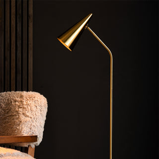 Lifestyle image of the Rockett St George Contemporary Brass Floor Lamp displayed in a dark room styled with an armchair. 