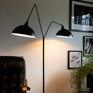 The Matt Black Double Floor Lamp displayed in a living room with a desk chair, art prints and a plant.