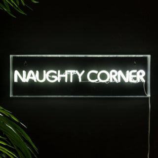 A neon sign that says 'naughty corner' styled on a black wall with some greenery.
