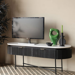 The Reeded Black Wood & Marble Low Console Table / TV Unit displayed with a screen and various decorative accessories on the top, on a geometric floor in front of a neutral wall. There is a plant to the left of the shot.