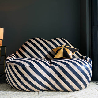 A striped sofa in the shape of some lips, styled on a rug in front of a dark wall with a cushion and a table lamp on a table.