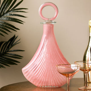 The Pink Glass Art Deco Decanter displayed with two cocktail glasses and a bottle on a metal table, with a plant in the background.