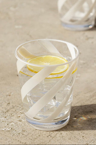 A beautiful water glass with cream patterned lines on the outside. The water glass tumbler is filled with water, ice cubes and lemon on an al fresco dining table.