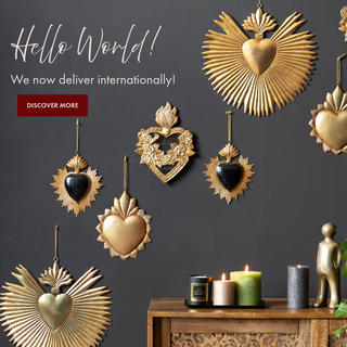 A square image of multiple gold metal milagro hearts on the wall. At the side is a sideboard styled with green candles and a human ornament. The text reads: Hello World! We now deliver internationally! Discover more.