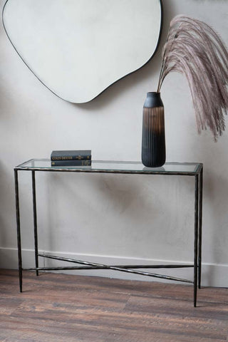 Slim console table with black metal legs and a glass top, featuring a cross-shaped support, styled with pampas grass and a book, below a statement mirror