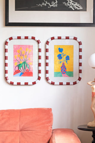 The Set of 2 Red & White Striped Frames with Colourful Flower Prints styled on the wall with other artwork, above a sofa, lamp and side table.