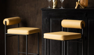 Two of the Sand Faux Leather Roll Back Bar Stools displayed in front of a black cabinet, styled with cocktail glasses and other home accessories.