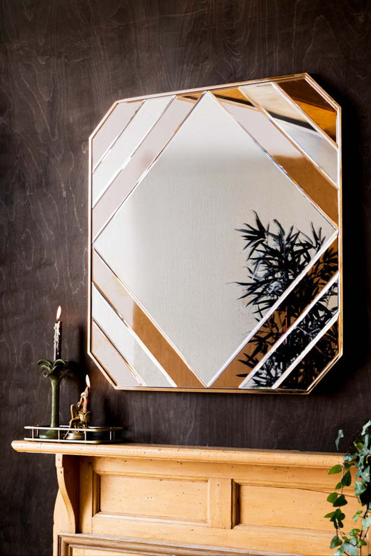 A square wall mirror styled on a dark wall above a fireplace, framed with lit candles, a trinket tray and a plant.