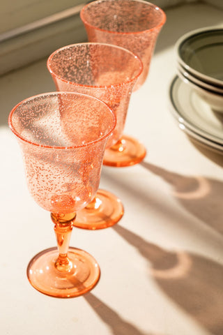 A lifestyle image of three acrylic coral pink picnic wine glasses lined up on a white surface.