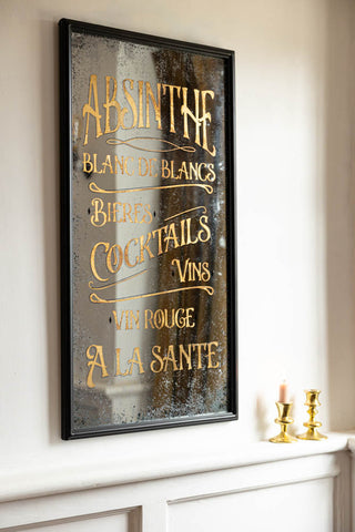 The Aged Style Absinthe, Bieres & Vins Decorative Mirror styled on a white wall, next to some gold candlestick holders.