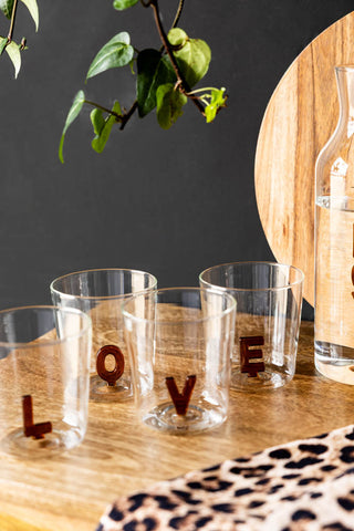 Clear glasses with 'LOVE' letters on a wooden surface with a dark background and plant.