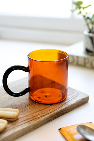 The Amber & Black Glass Mug on a wooden board, styled with some biscuits, a plant and some accessories.