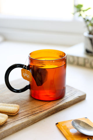 The Amber & Black Glass Mug with a teabag and liquid inside, styled on a wooden board with various accessories and some biscuits.
