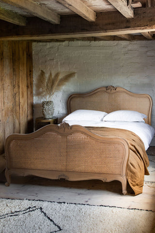 The Antique-Style Wooden Bedframe with pillow, a duvet and throw on, next to a side table with some dried grasses in a vase.