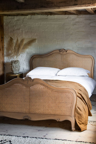The Antique-Style Wooden Bedframe  in a bedroom with a duvet and pillows on, styled with a side table and dried grasses in a vase.