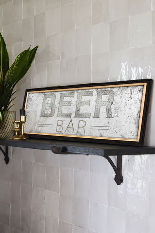The Vintage-Style Beer Bar decorative Mirrored wall art displayed on a shelf on a tiled wall, with some candlestick holders and a potted plant.