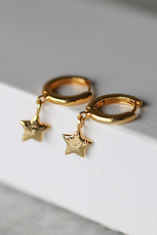 Lifestyle image of the You're A Star Gold Huggie Hoop Earrings displayed draped over the corner of a white surface. 