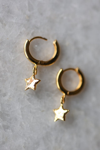 Lifestyle image of the You're A Star Gold Huggie Hoop Earrings displayed on a white marble surface. 