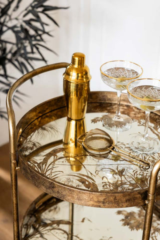 Gold cocktail shaker and glasses on a decorative glass cart.
