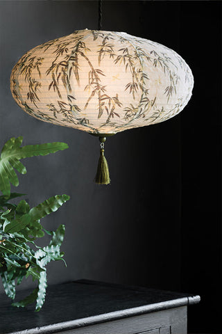 A textured ceiling lampshade with a green tassel, featuring a bamboo pattern in front of a dark background, styled with a plant on a sideboard.