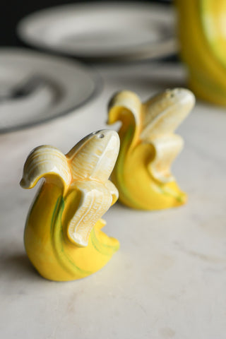 The Banana Salt & Pepper Shakers displayed on a marble table with other kitchen accessories in the background.