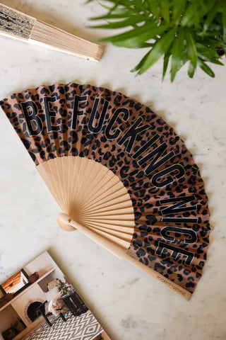 The Be Fucking Nice Leopard Print Fan displayed on a white marble table with another fan, magazine and a plant.