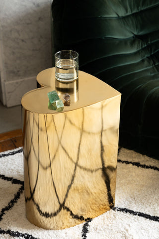 The Beautiful Brass Heart Side Table with a glass of water and some crystals on, styled on a rug next to a sofa.