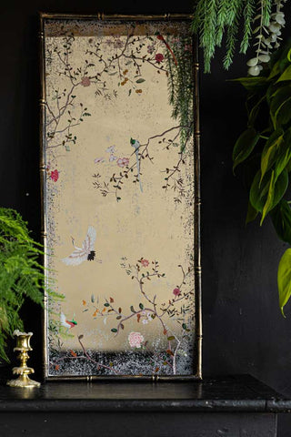 The Beautiful Pink Chinoiserie Decorative Mirrored wall art styled leaning against the wall on a black sideboard, with a candlestick holder and some greenery.