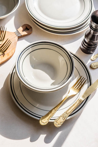 A lifestyle image of the bistro style black and white dinnerware on a white table with a gold knife and fork placed on the plate.