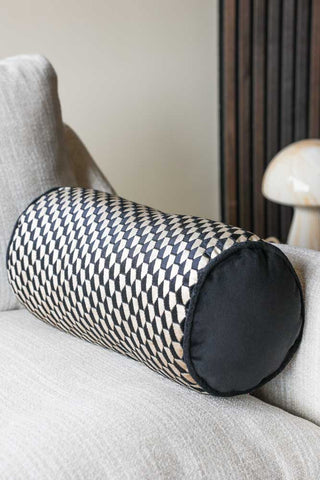 Lifestyle image of the Black Geo Bolster Cushion displayed on a sofa with a lamp in the background. 