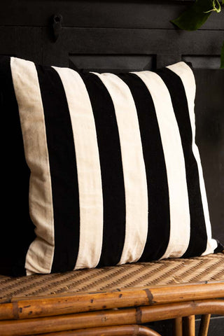 The Black & Off-White Stripe Velvet Cushion displayed on a wicker bench in front of a black sideboard.