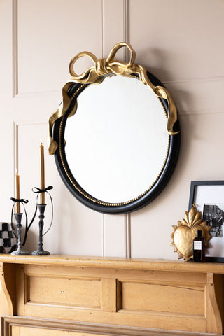 The Black & Gold Bow Round Wall Mirror styled on a panelled wall, with various decorative accessories surrounding it.