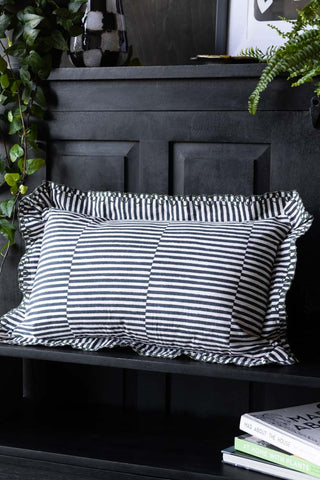 The Black & White Stripe Cushion With Scalloped Edge on a bench in front of a sideboard, styled with some books, plants and other accessories.
