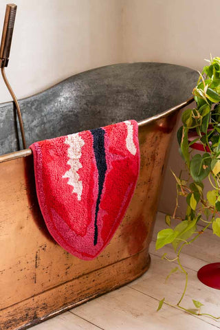 Red lip-shaped bath mat draped over a metal bath, styled next to a plant on a table.