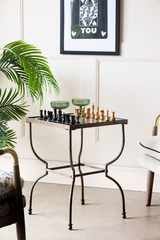 The Bronze Effect Chess Side Table with chess pieces and 2 glasses on it, styled with a armchair and plant in the background.