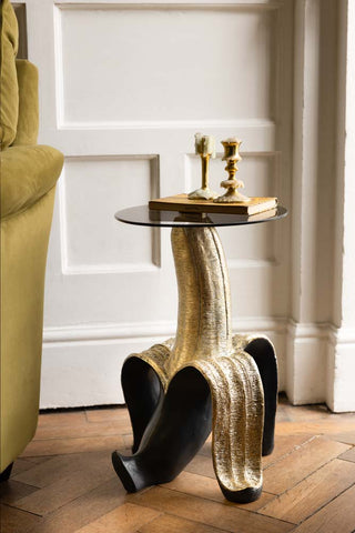 The Charcoal & Gold Banana Side Table displayed in a living room next to a sofa, with a book and candlesticks on top.