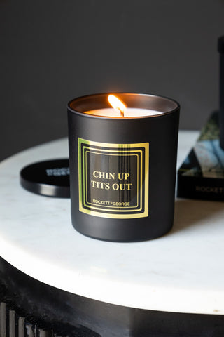 The Chin Up, Tits Out Scented Candle lit and displayed on a white surface, with the lid and a book in the background.