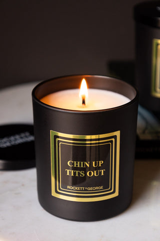 Close-up of the lit Chin Up, Tits Out Scented Candle on a white surface.