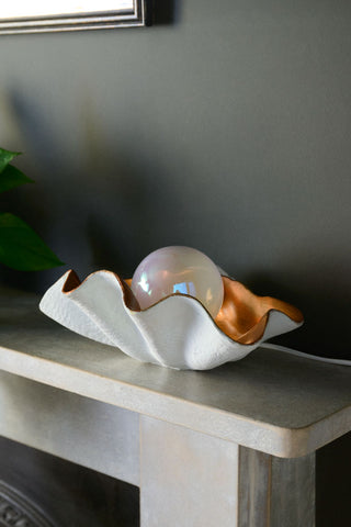 The Clam Table Lamp displayed on a fireplace shelf next to a plant.