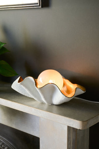 The Clam Table Lamp displayed on a mantelpiece with a plant in the background.