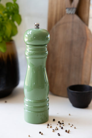 The Classic Pepper Mill in Green on a work surface with some pepper, plant, wooden boards and a black pot.