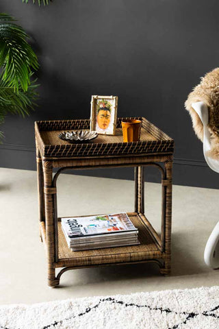 Lifestyle image of the Dark Brown Rattan Side Table styled with various home accessories and magazines, next to a chair, plant and rug in front of a black wall.